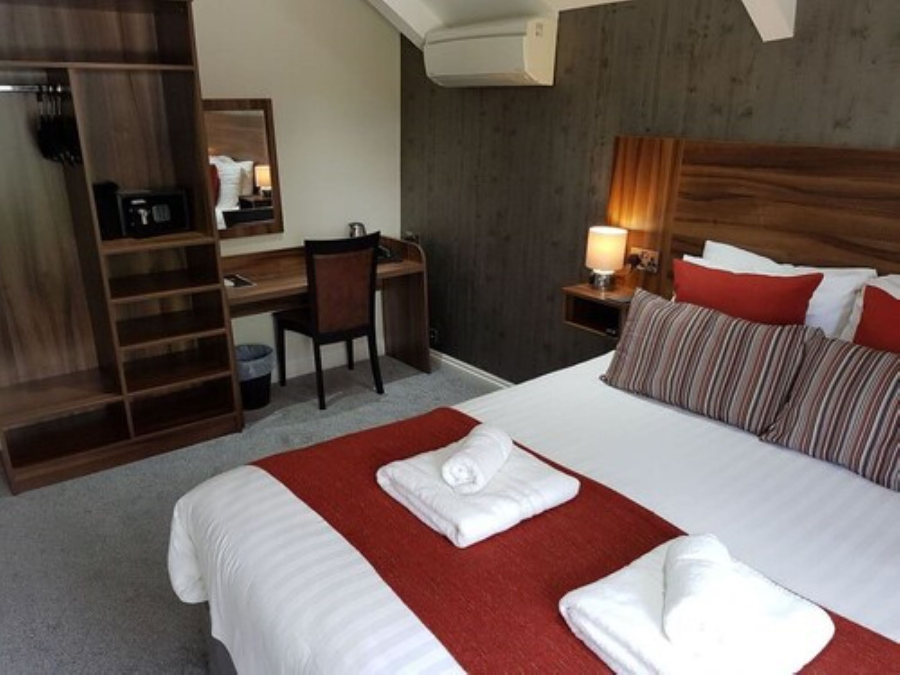 Una habitación de hotel con cama, escritorio, silla, armario y mesita de noche con lámpara.