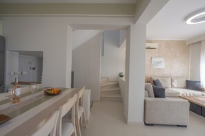 A modern living room with a sofa, a dining area with a table and chairs, and a staircase leading to another level. - Valea Villa (Naxos)