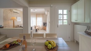 A dining area with a table set for two, featuring a fruit platter, a bottle of wine, and glasses. There is a view into the adjacent living room through an open door. - Valea Villa (Naxos)