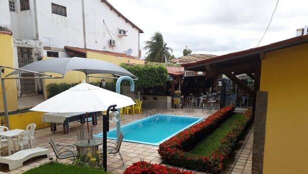 Una piscina con sillones de descanso y sombrillas, rodeada por un jardín con flores rojas y un edificio amarillo al fondo.