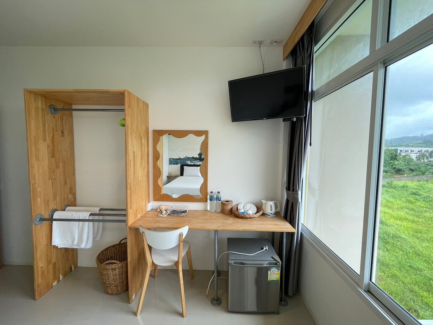 Habitación de hotel compacta con escritorio de madera, una silla, un cubo de basura pequeño, televisor montado en la pared y un ventanal con vistas al exterior.