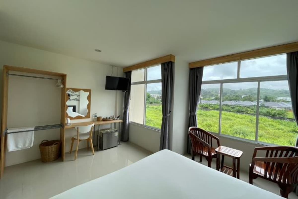 A hotel room with a bed, a desk with a chair, a TV, and large windows with a view of greenery.