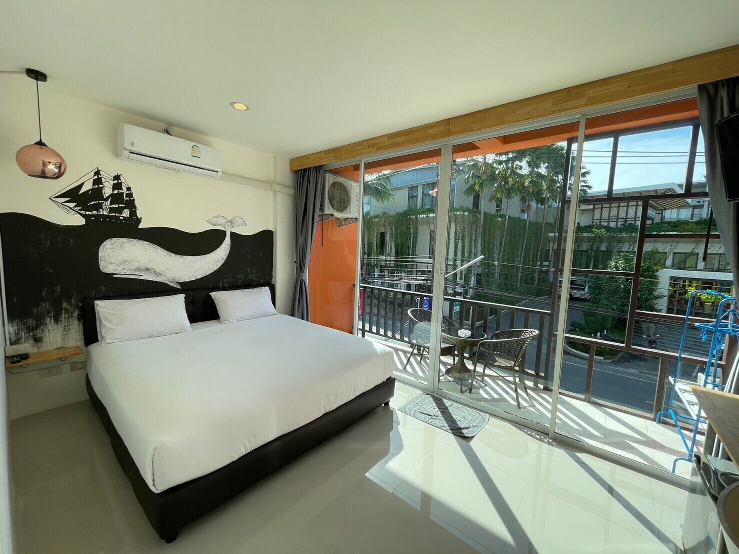 Une chambre d’hôtel moderne dotée d’un grand lit, offrant une vue sur la ville grâce à de larges fenêtres du sol au plafond, et disposant d’un balcon avec mobilier d’extérieur.