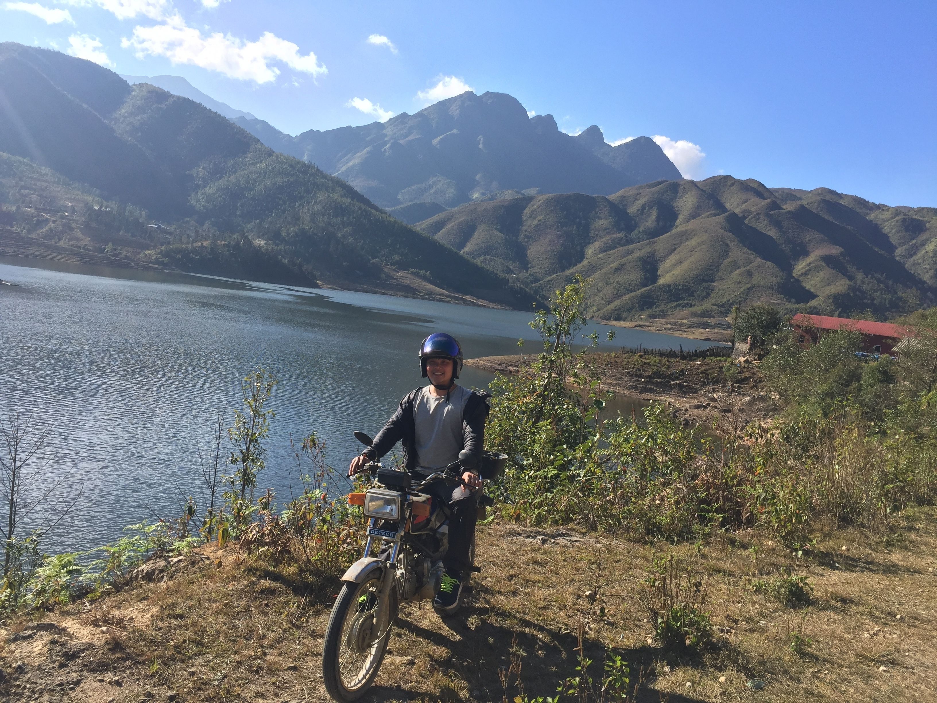 Una persona en motocicleta cerca de un lago con montañas al fondo.