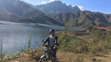 Una persona en motocicleta cerca de un lago con montañas al fondo.