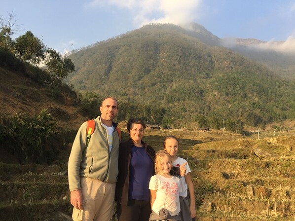 Une famille se tient dans une région montagneuse, avec des champs en terrasses et des maisons traditionnelles en arrière-plan.