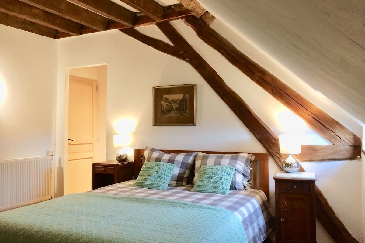 Um quarto com teto inclinado, uma cama com colcha xadrez, duas luminárias de cabeceira, uma mesa de cabeceira de madeira e um quadro na parede.
