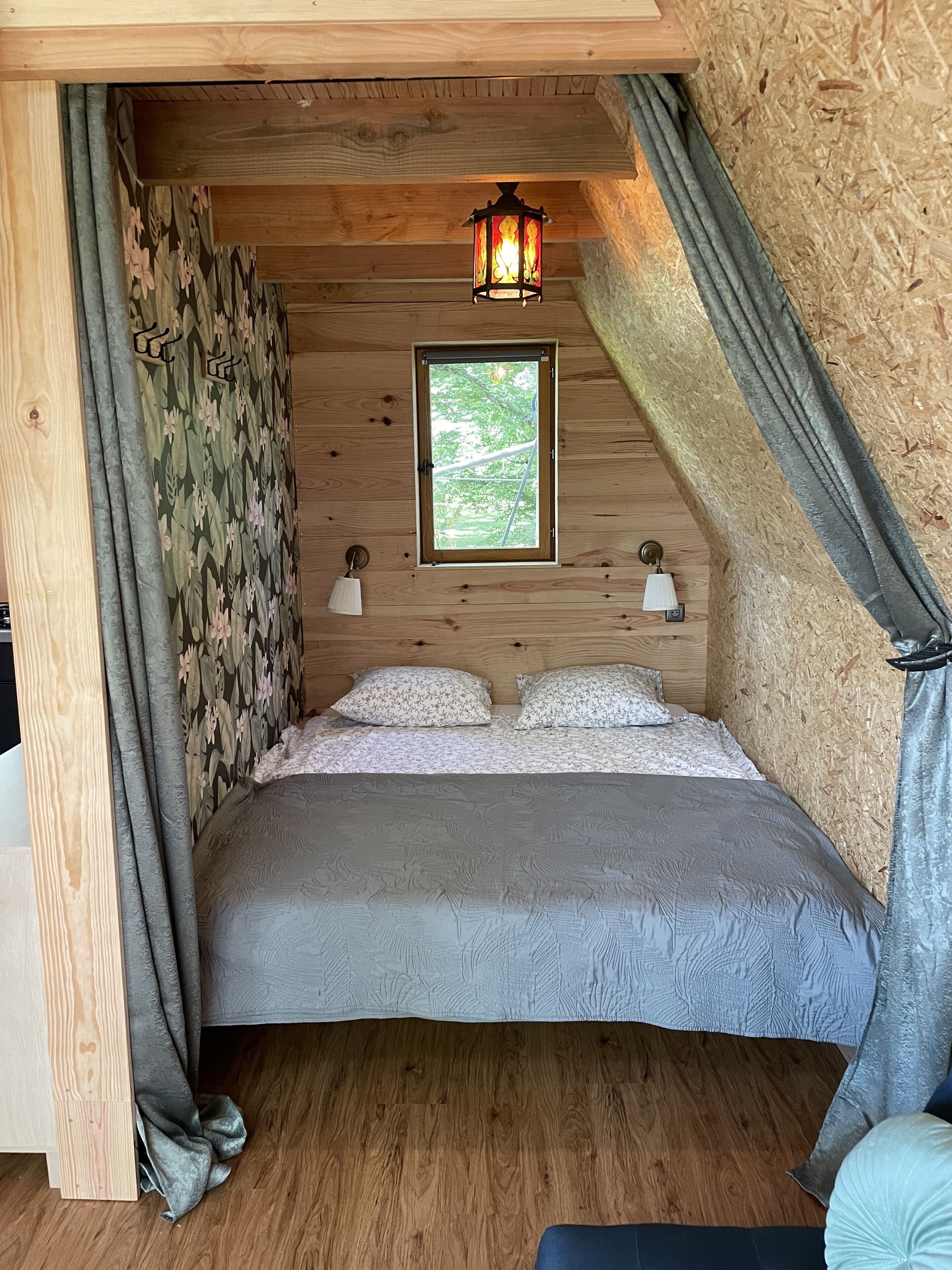 A cozy bedroom with a wooden ceiling, a window with a view of greenery, a bed with floral bedding, and two wall-mounted lamps.
