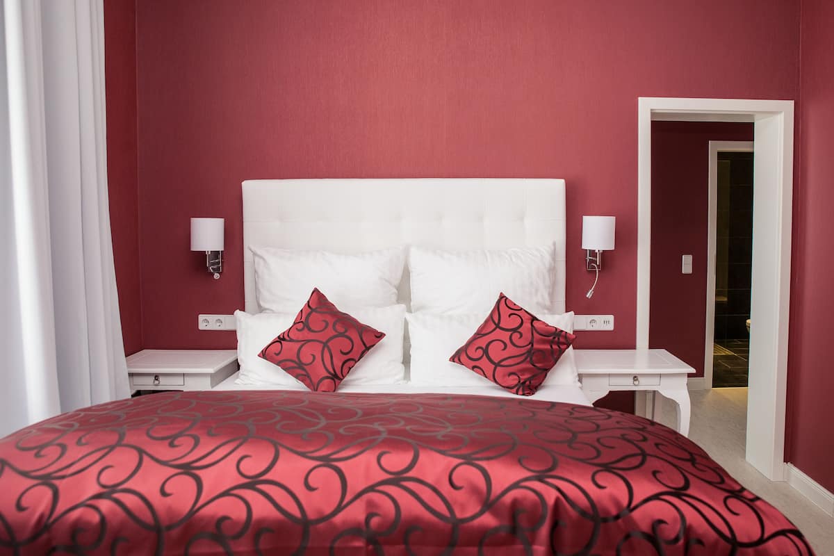 A neatly made bed with a red and black patterned bedspread, white headboard, and two red patterned pillows. There are two bedside tables with white lamps.