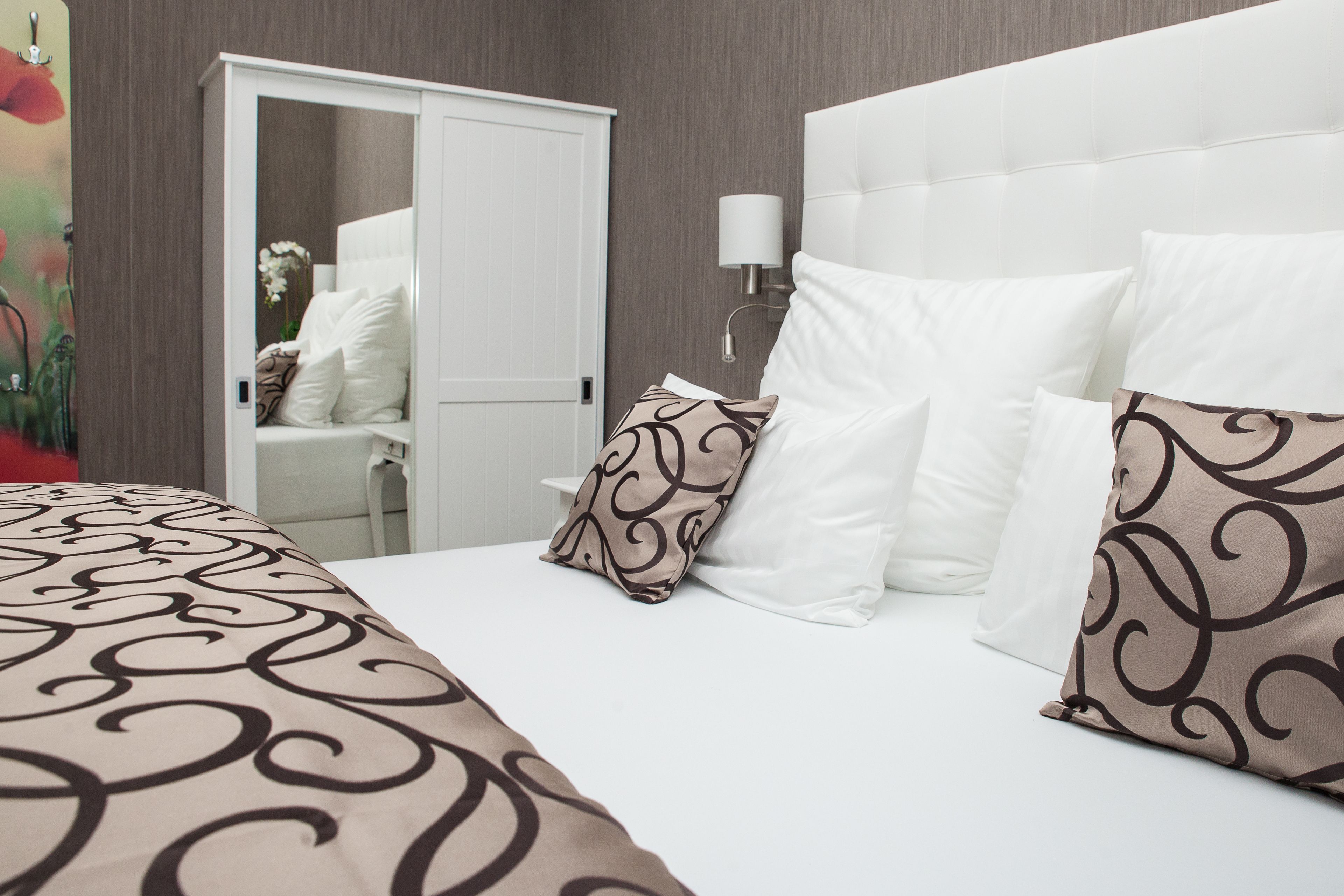 A hotel room with a large bed, white bedding, a white headboard, a bedside table with a lamp, and a wardrobe.