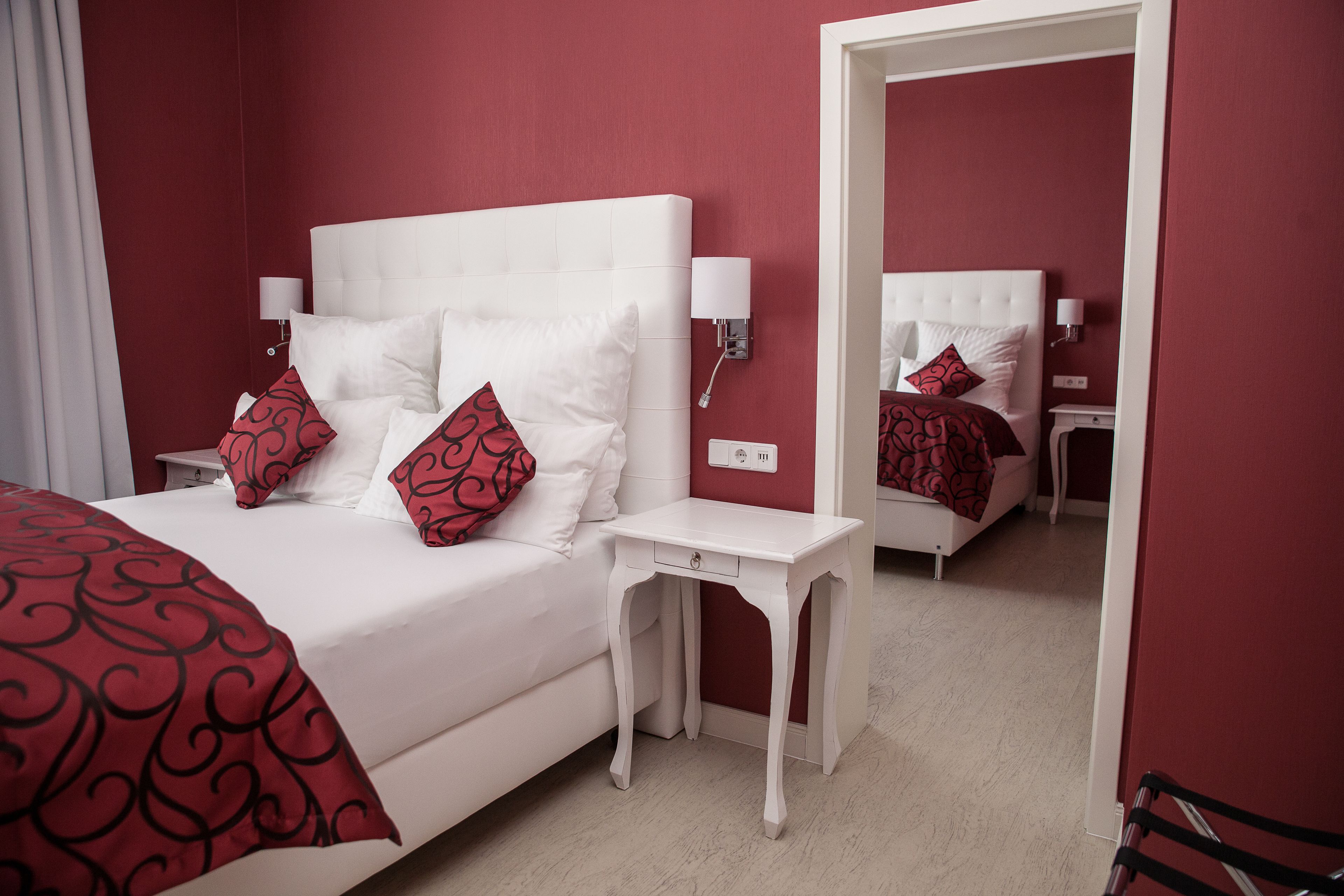 Una habitación de hotel con una cama blanca, almohadones estampados rojos, una mesita de noche blanca y una pared roja.