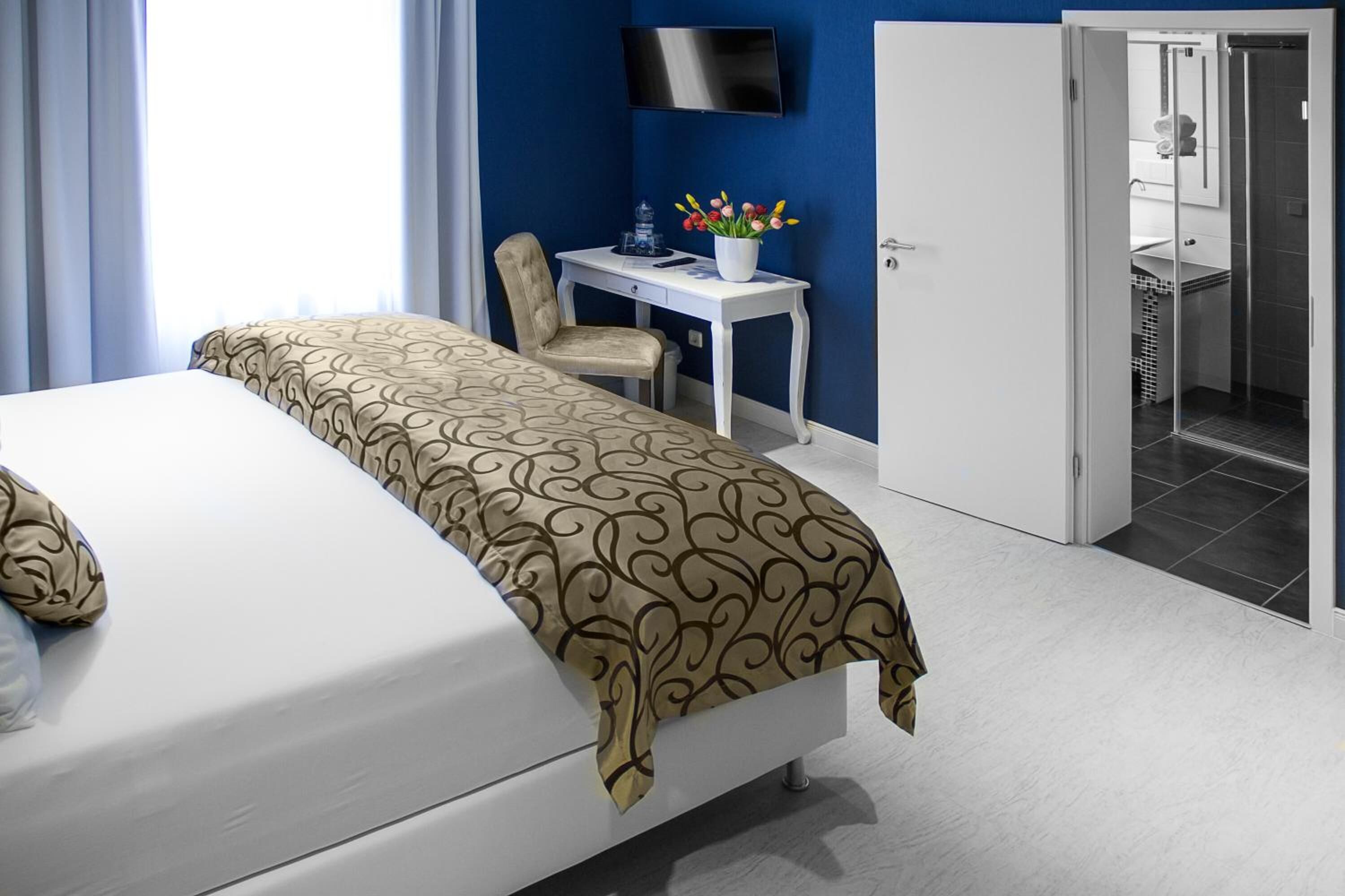 Ein Hotelzimmer mit einem Bett, einem Schreibtisch mit Stuhl, einem an der Wand montierten Fernseher und einem durch eine geöffnete Tür einsehbaren Badezimmer.