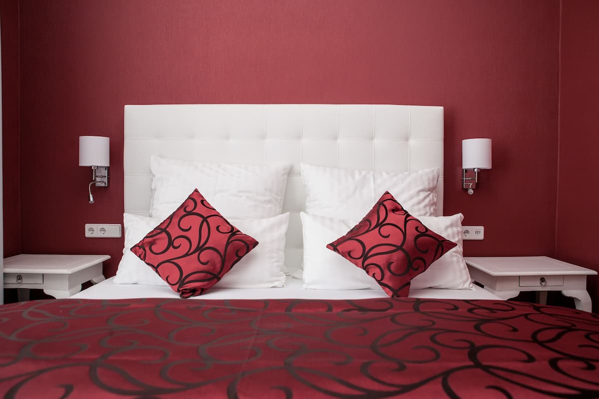 A hotel room with a white headboard, two bedside tables, a red patterned bedspread, and red and white pillows.