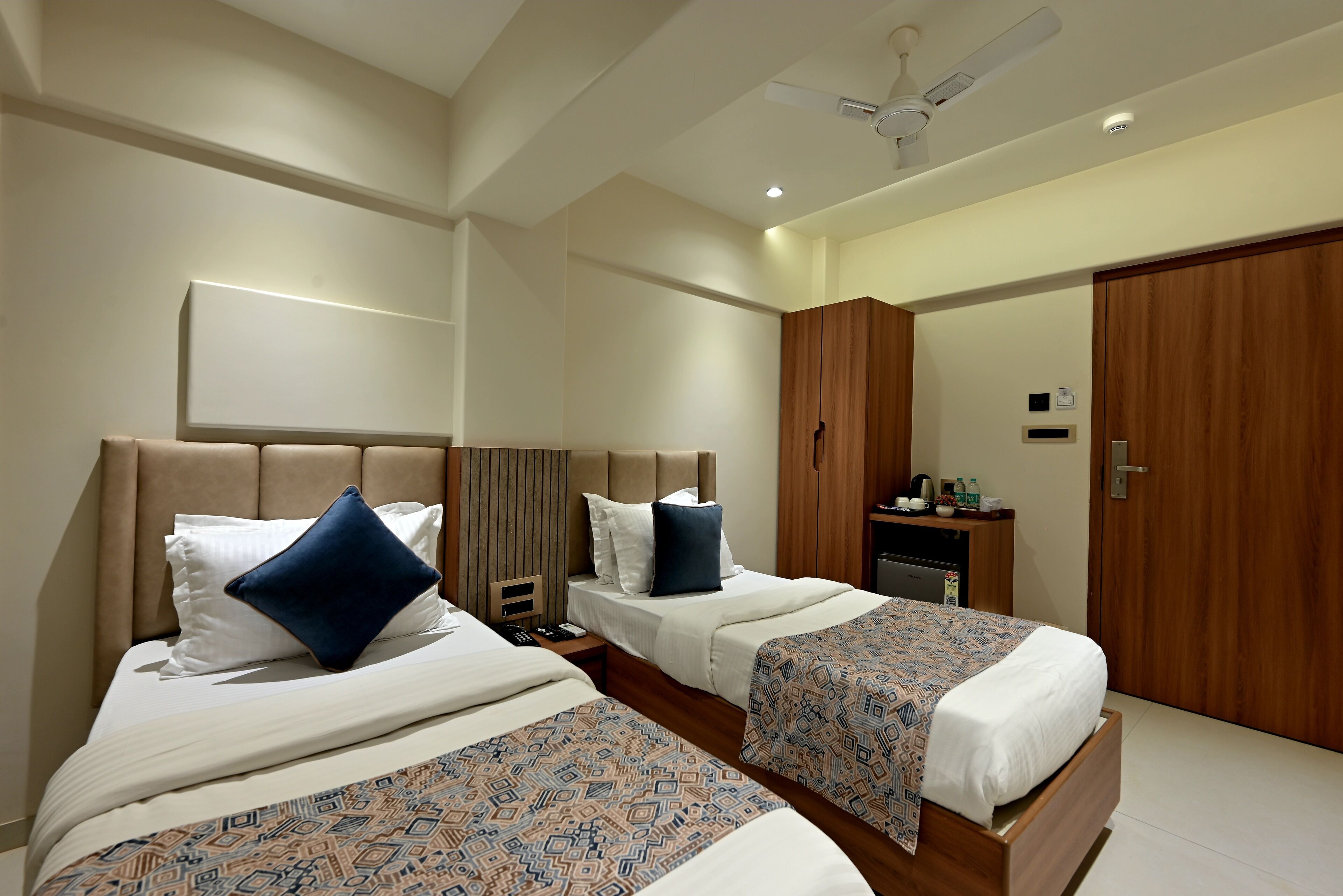 A hotel room with two beds, a ceiling fan, a minibar, and a wooden wardrobe.