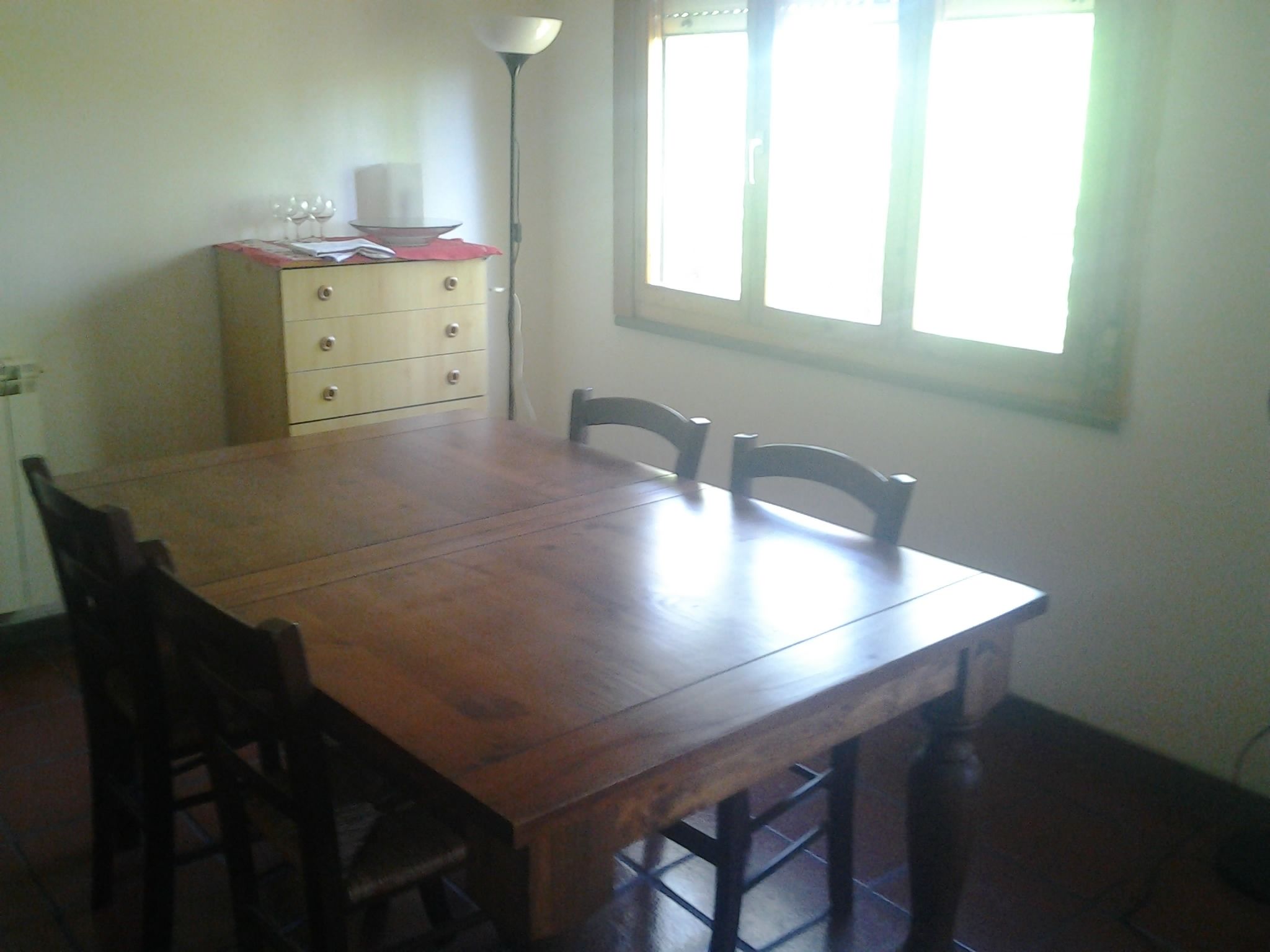 A dining area with a wooden table, four chairs, a small cabinet with drawers, and a floor lamp.