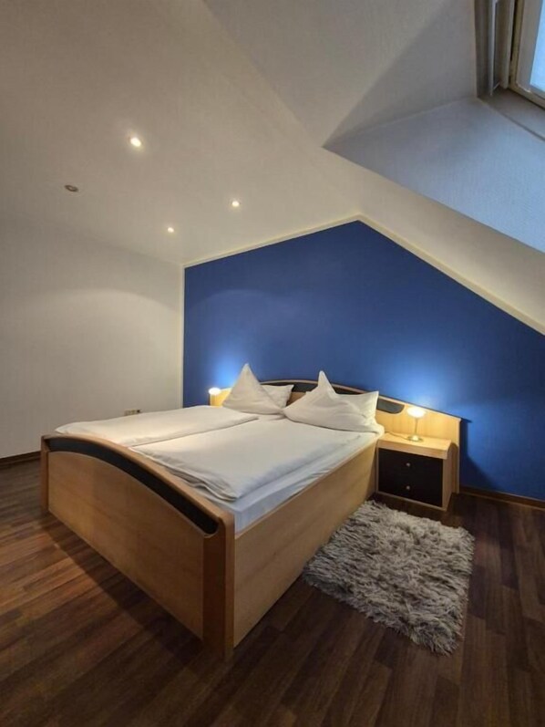 A bedroom with a sloped ceiling, a bed with white bedding, a nightstand with a lamp, and a fluffy rug.
