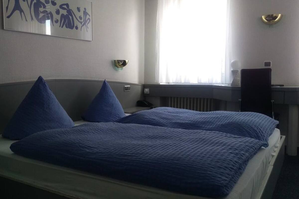 A hotel room with two beds, a window with blinds, a radiator, and a desk with a lamp.