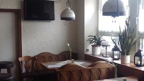 Un área de comedor con mesa y sillas de madera, una lámpara colgante, un televisor en la pared y una ventana con plantas.
