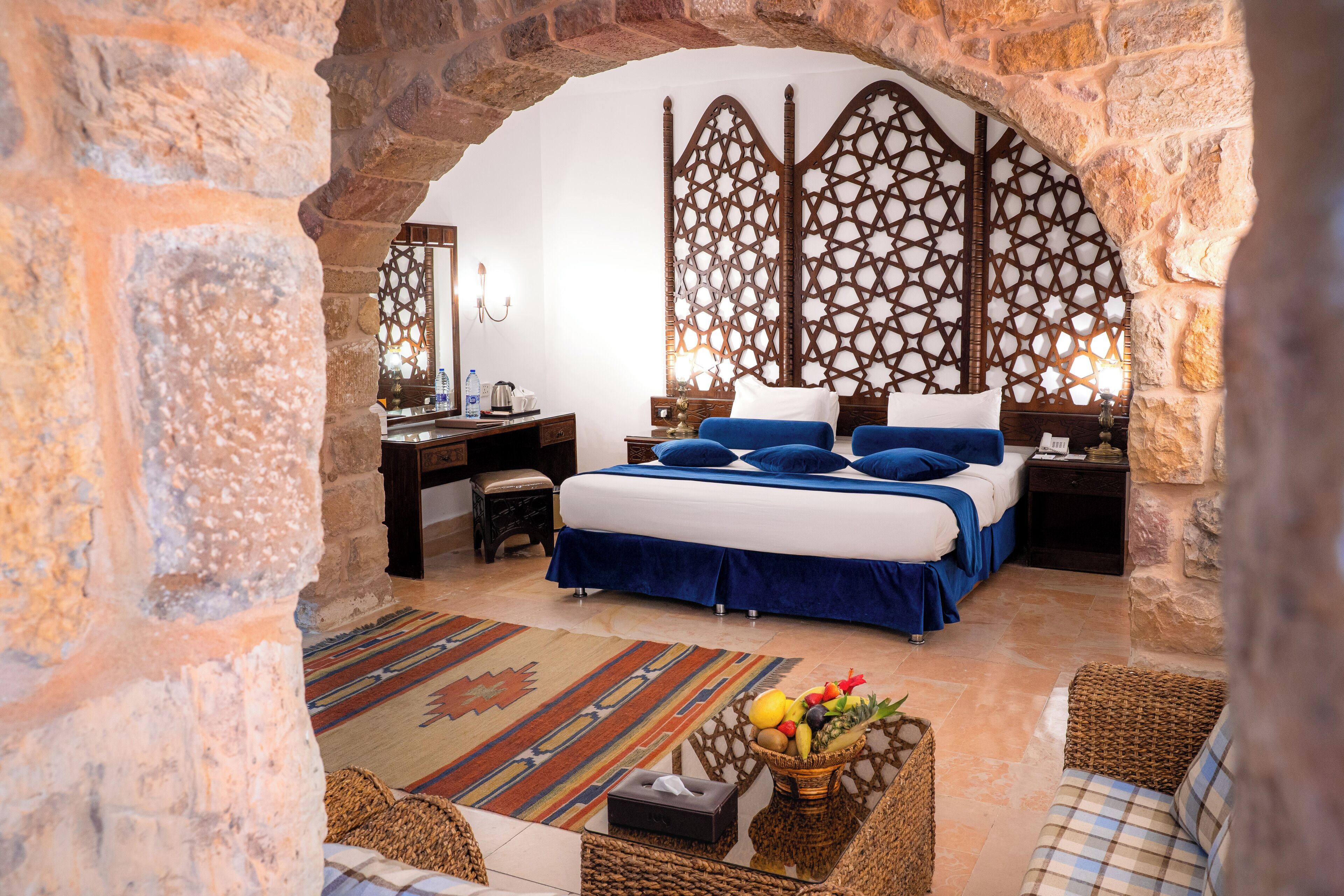 A bedroom with a stone archway, a bed with blue bedding, a nightstand with a lamp, and a woven basket with fruits.
