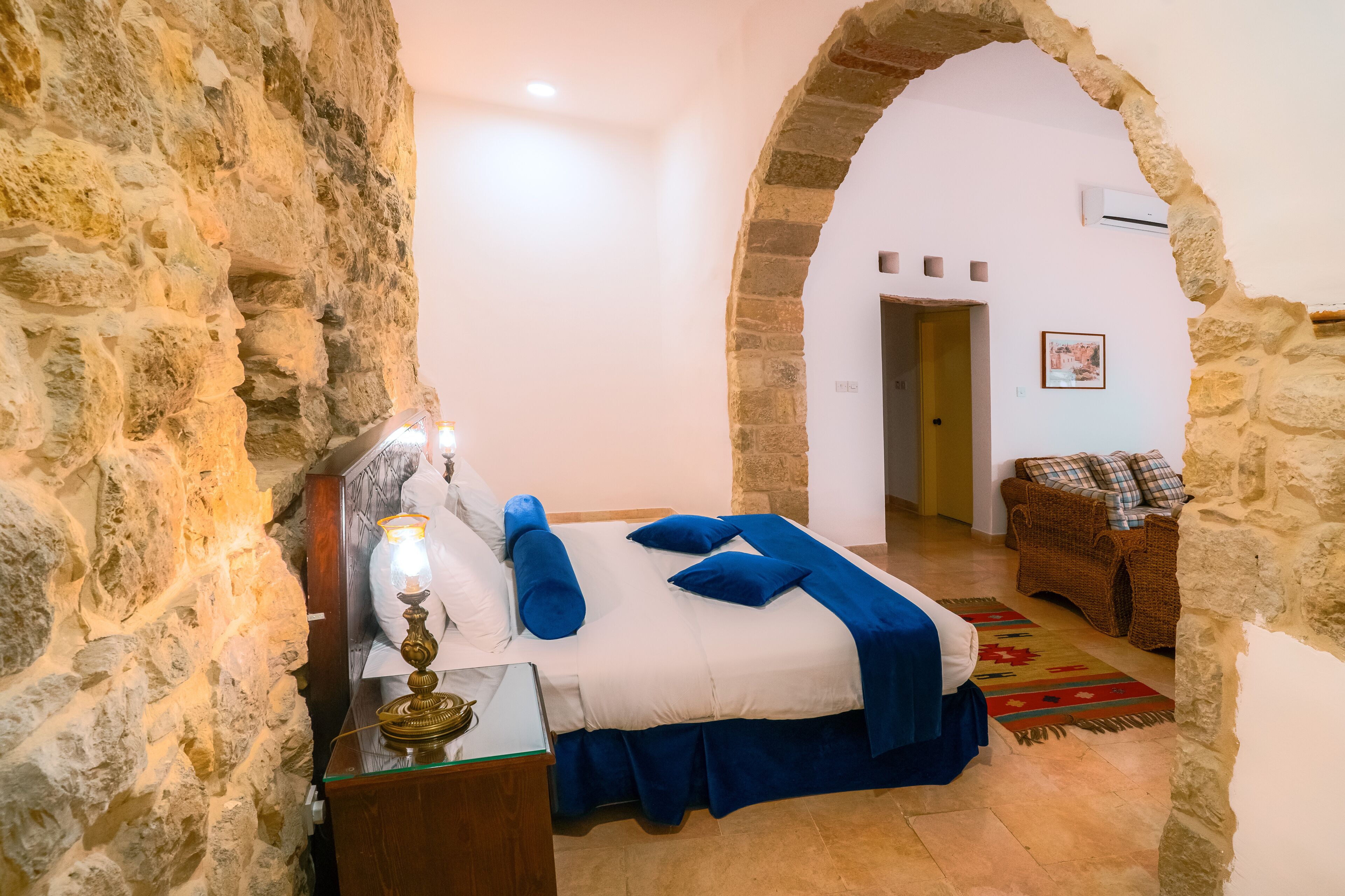 Quarto com parede de pedra, cama com roupa de cama azul, mesa de cabeceira com abajur e uma cadeira de vime.