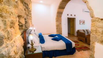 Quarto com parede de pedra, cama com roupa de cama azul, mesa de cabeceira com abajur e uma cadeira de vime.