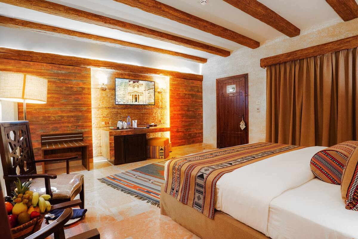 A hotel room with a wooden ceiling, a bed with patterned bedding, a bench, a fruit basket, and a wall-mounted television.