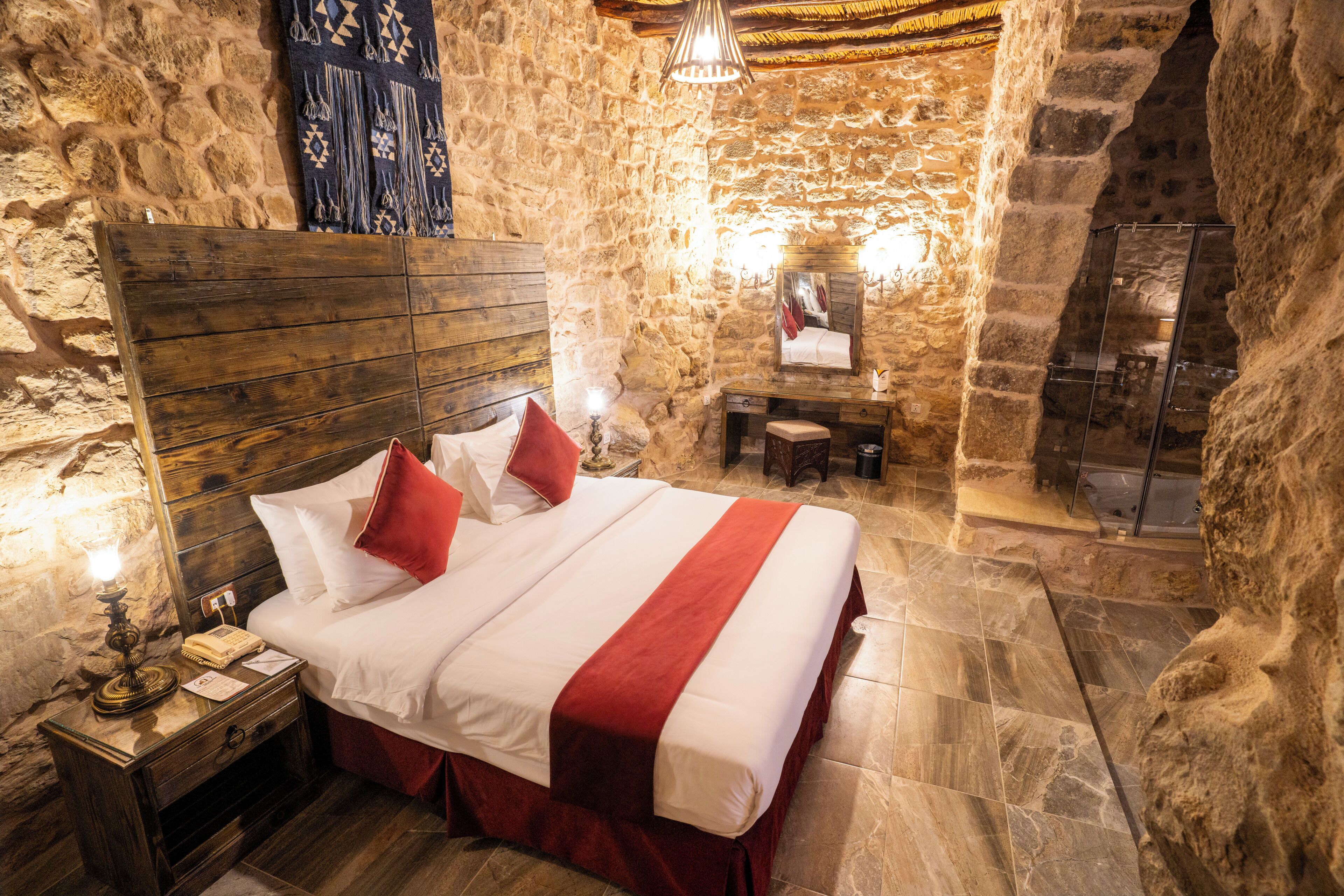 A stone-walled room with a wooden bed, a nightstand, a small table, and a mirror.