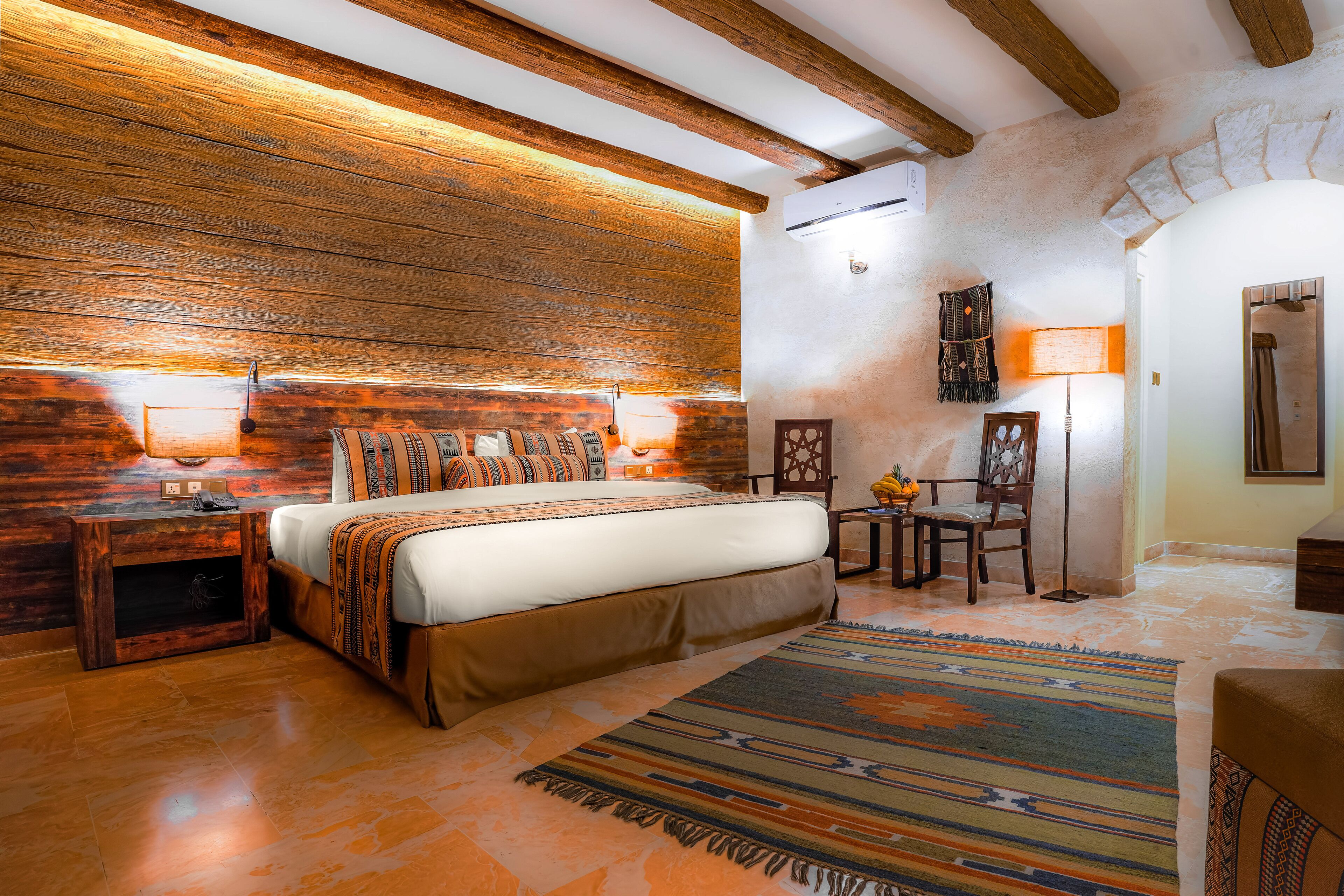 A bedroom with a wooden headboard, a bed with patterned bedding, a nightstand, a lamp, and a chair.