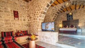 A stone-walled room with a stone bench, a fruit basket, and a bed with a red bedspread. - Old Village Resort (Wadi Musa)