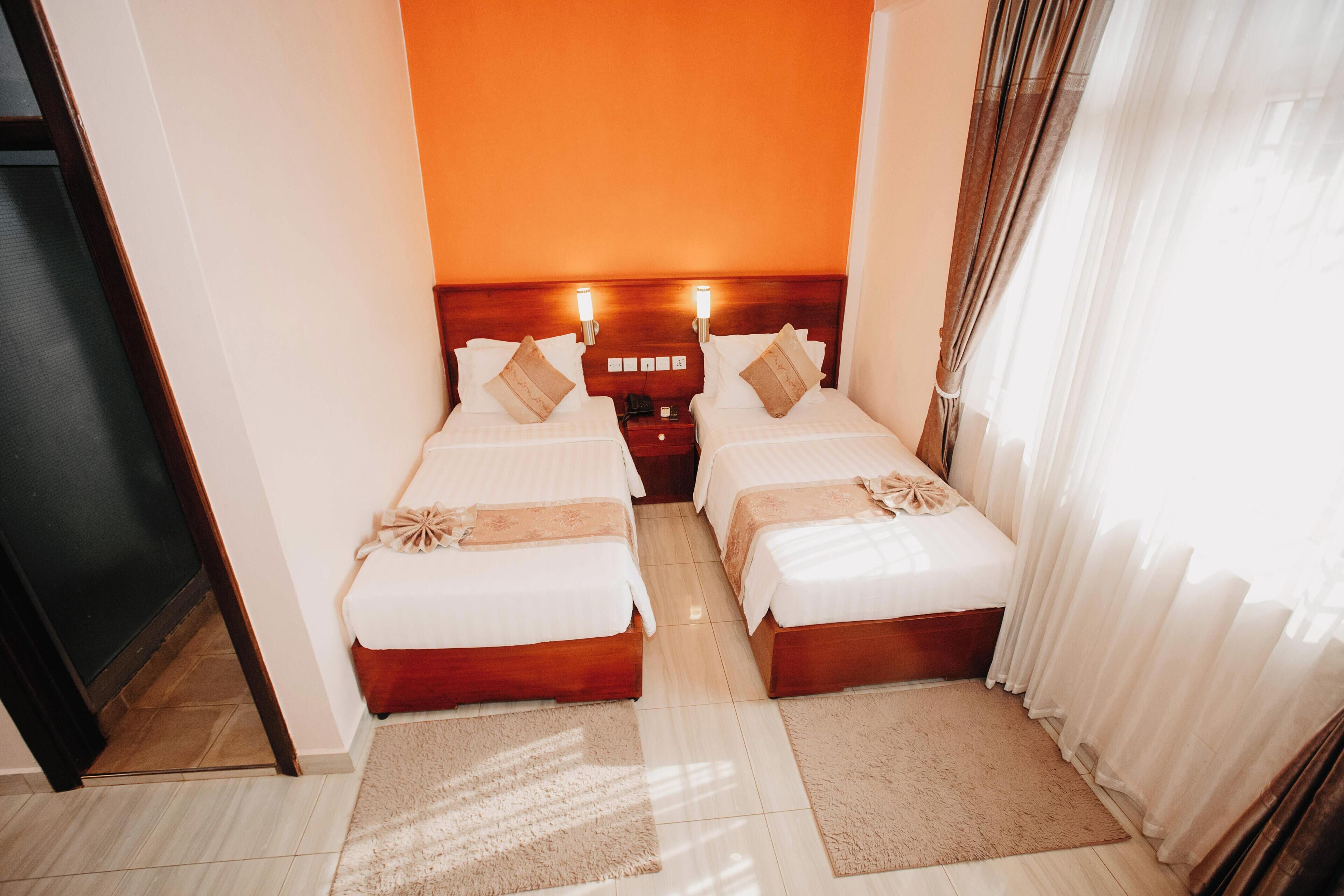 A hotel room with two beds, a wooden headboard, a nightstand, and a large window with sheer curtains.