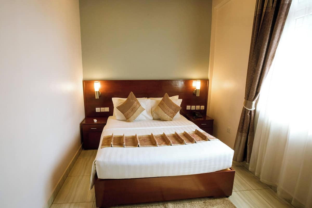 Una cama bien hecha con cabecera de madera, dos mesitas de noche con lámparas y una ventana con cortinas.