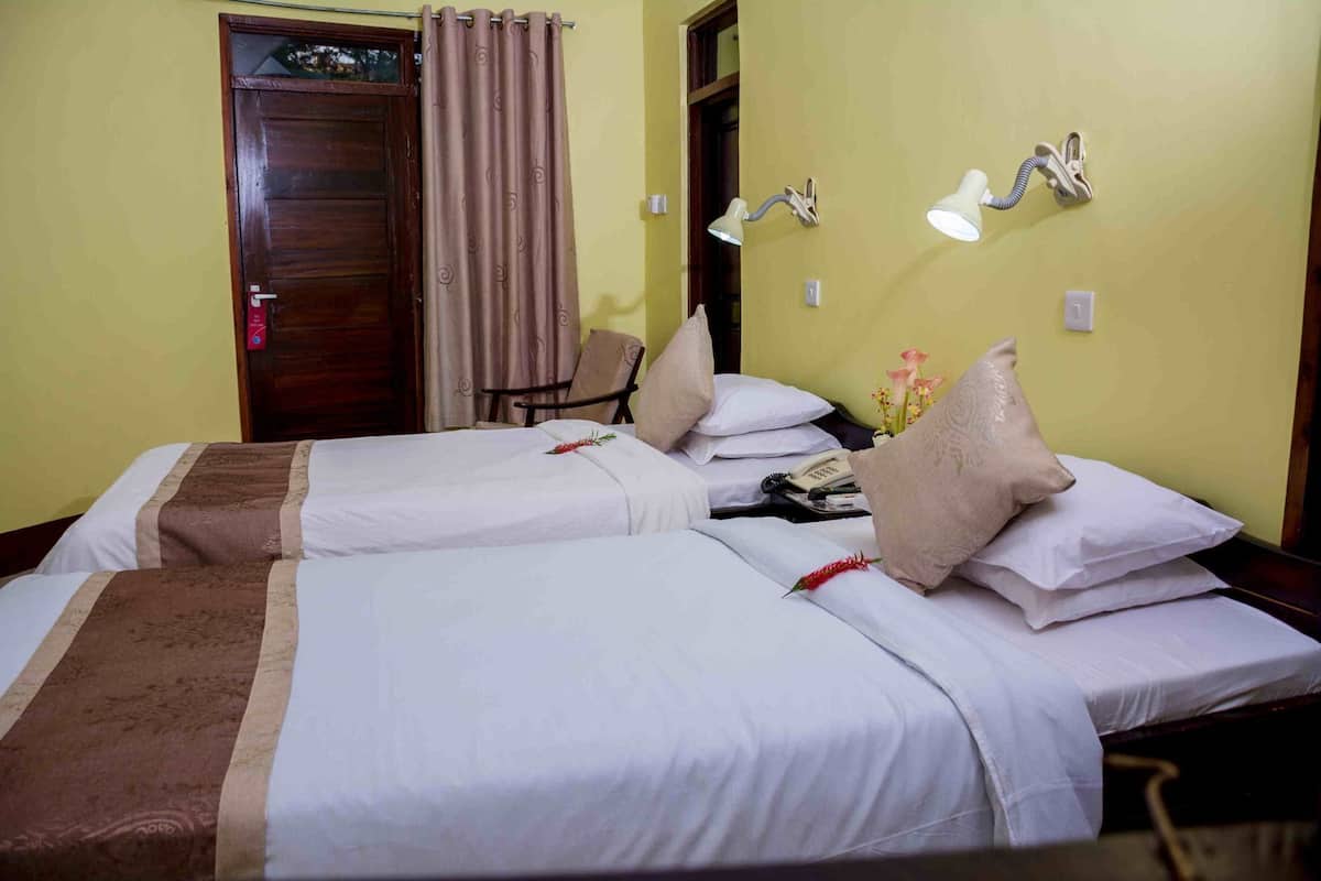 Habitación de hotel con dos camas, una puerta de madera, una cortina, una silla, una mesita de noche con teléfono y lámparas fijadas a la pared.
