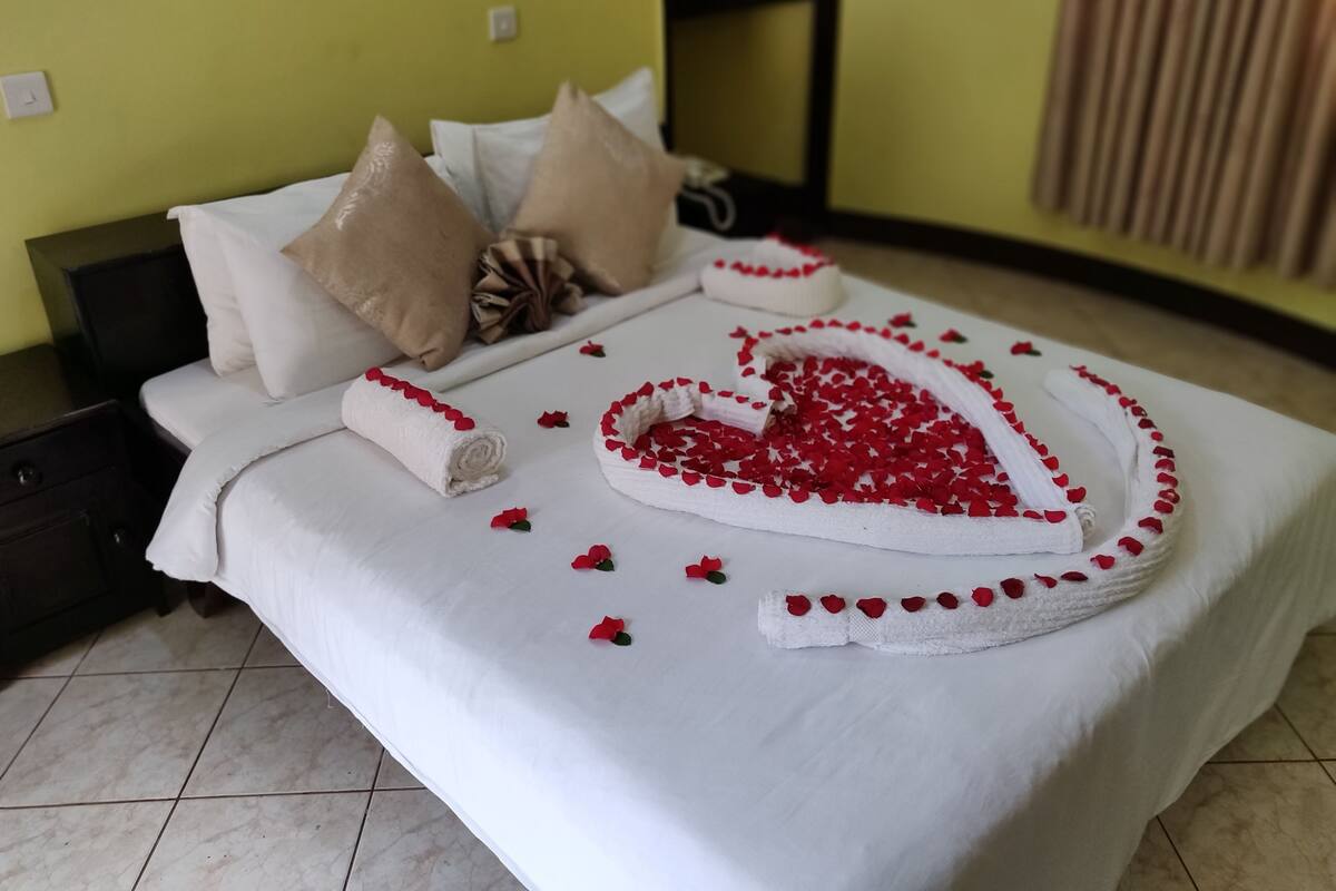 Una cama bien hecha con un arreglo de pétalos de rosa en forma de corazón y una toalla enrollada.