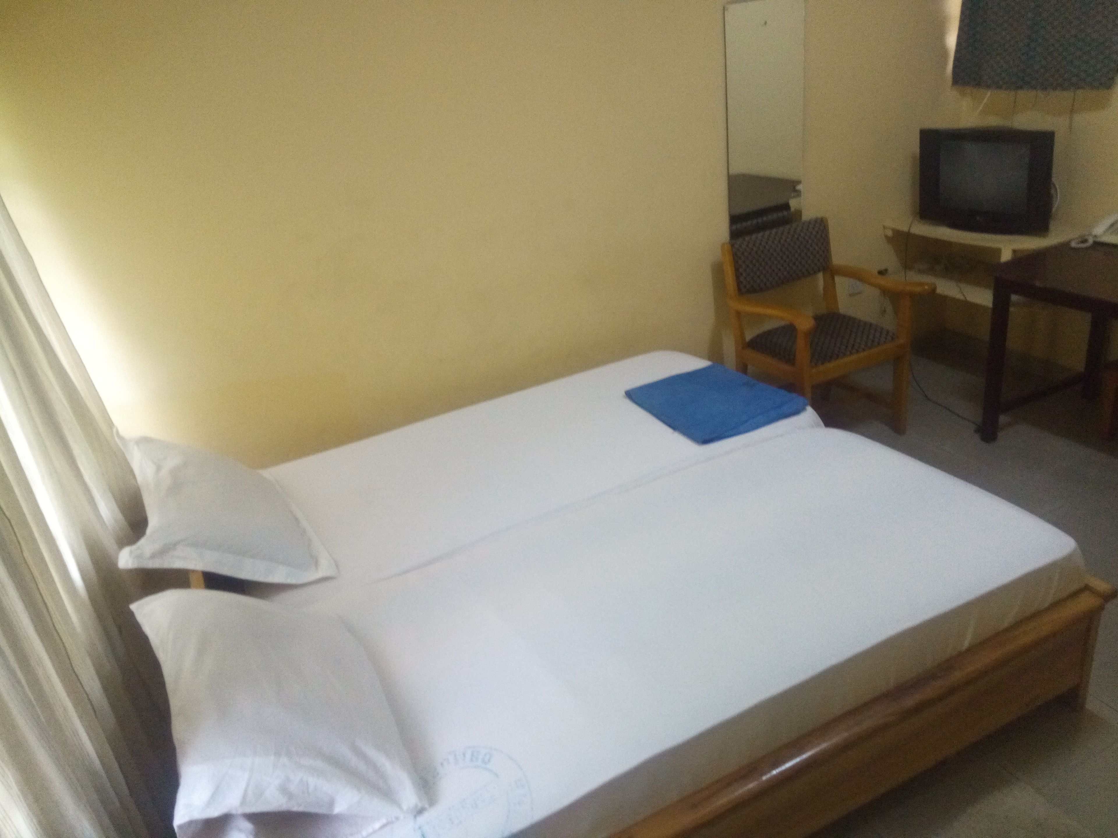 A single bed with white linens, a blue pillow, and a wooden headboard. A television is mounted on a wooden stand to the right of the bed.