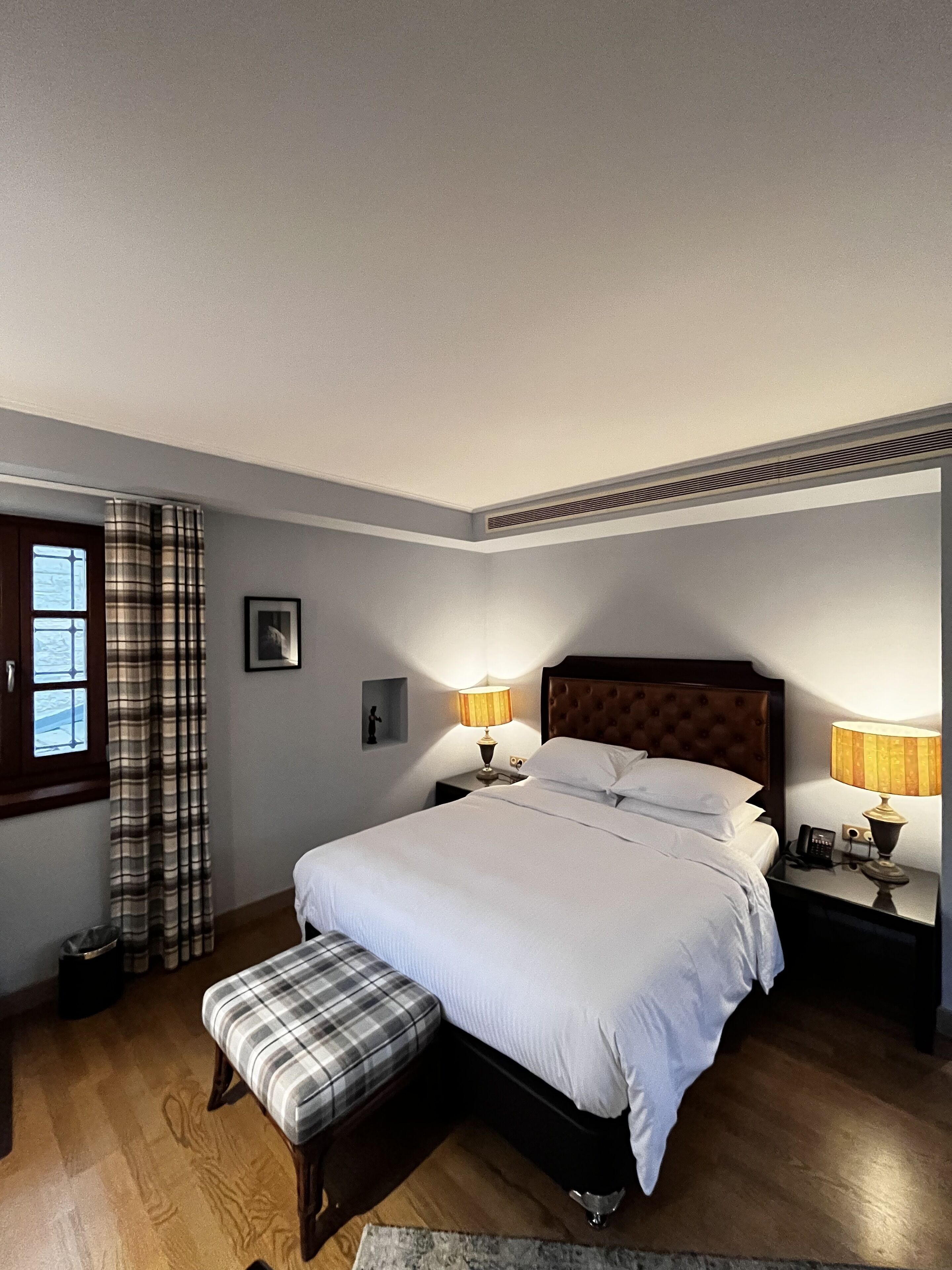 A neatly arranged bedroom with a large bed, two bedside tables with lamps, a bench, a window with curtains, and a framed picture on the wall.