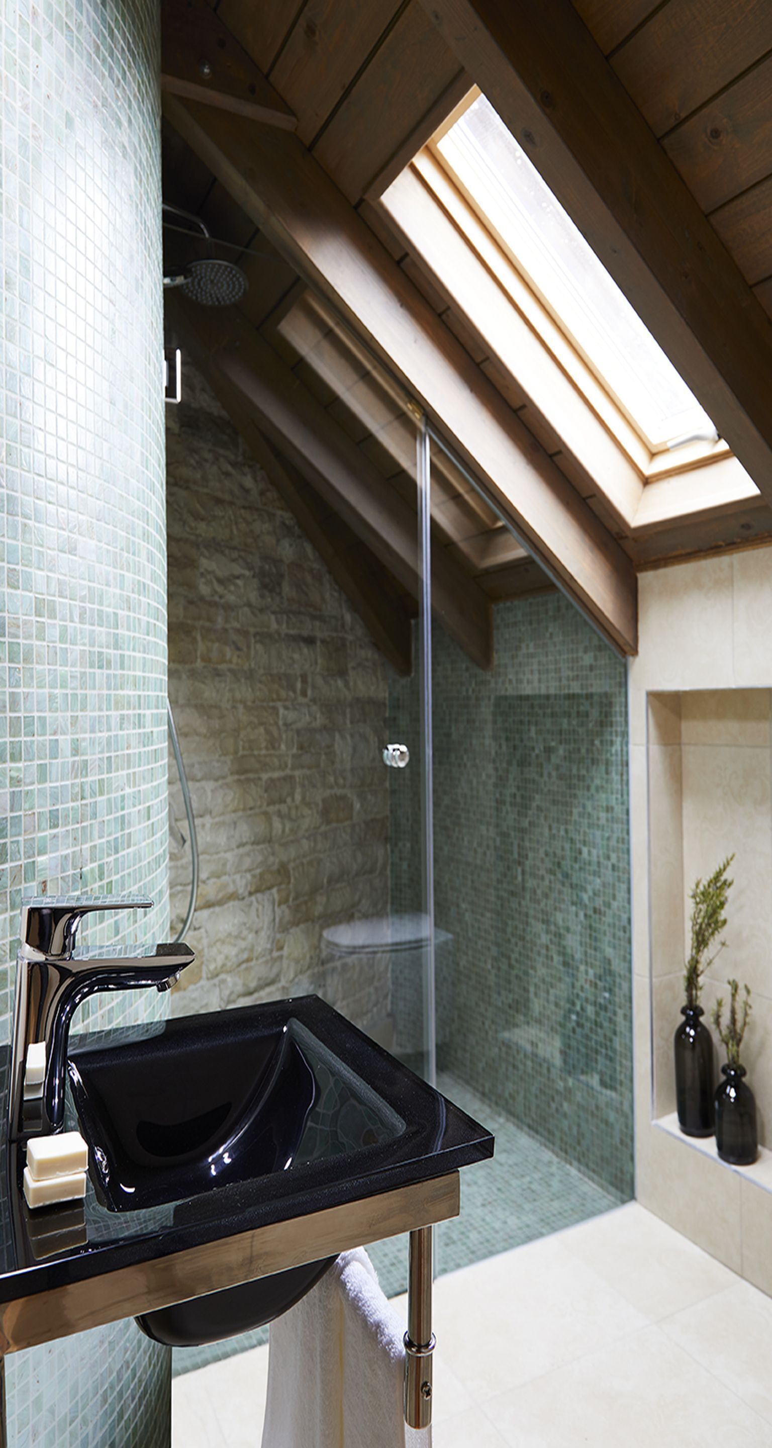 Een moderne badkamer met een douche met glazen wand, een zwart wastafel en een dakraam erboven.