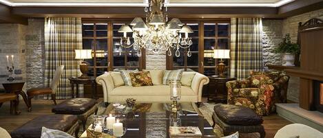 A spacious living room with a large sofa, armchairs, and a coffee table. There is a fireplace, a chandelier, and a stone wall.