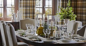 A dining table set with plates, glasses, and candles, with a window view and plaid curtains in the background. - MounTrace Suites & Spa (Zagori)