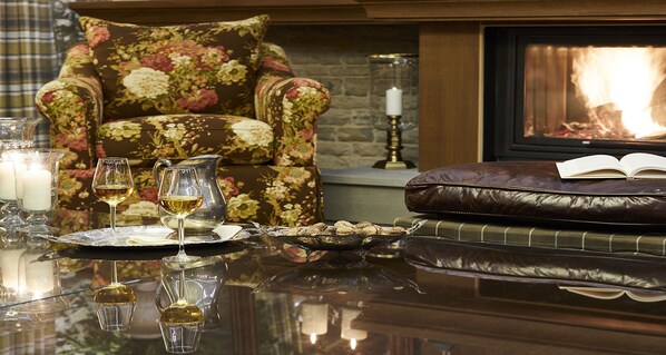 A cozy living room with a fireplace, a floral armchair, a leather ottoman, and a glass table with wine glasses and a candle. - MounTrace Suites & Spa (Zagori)