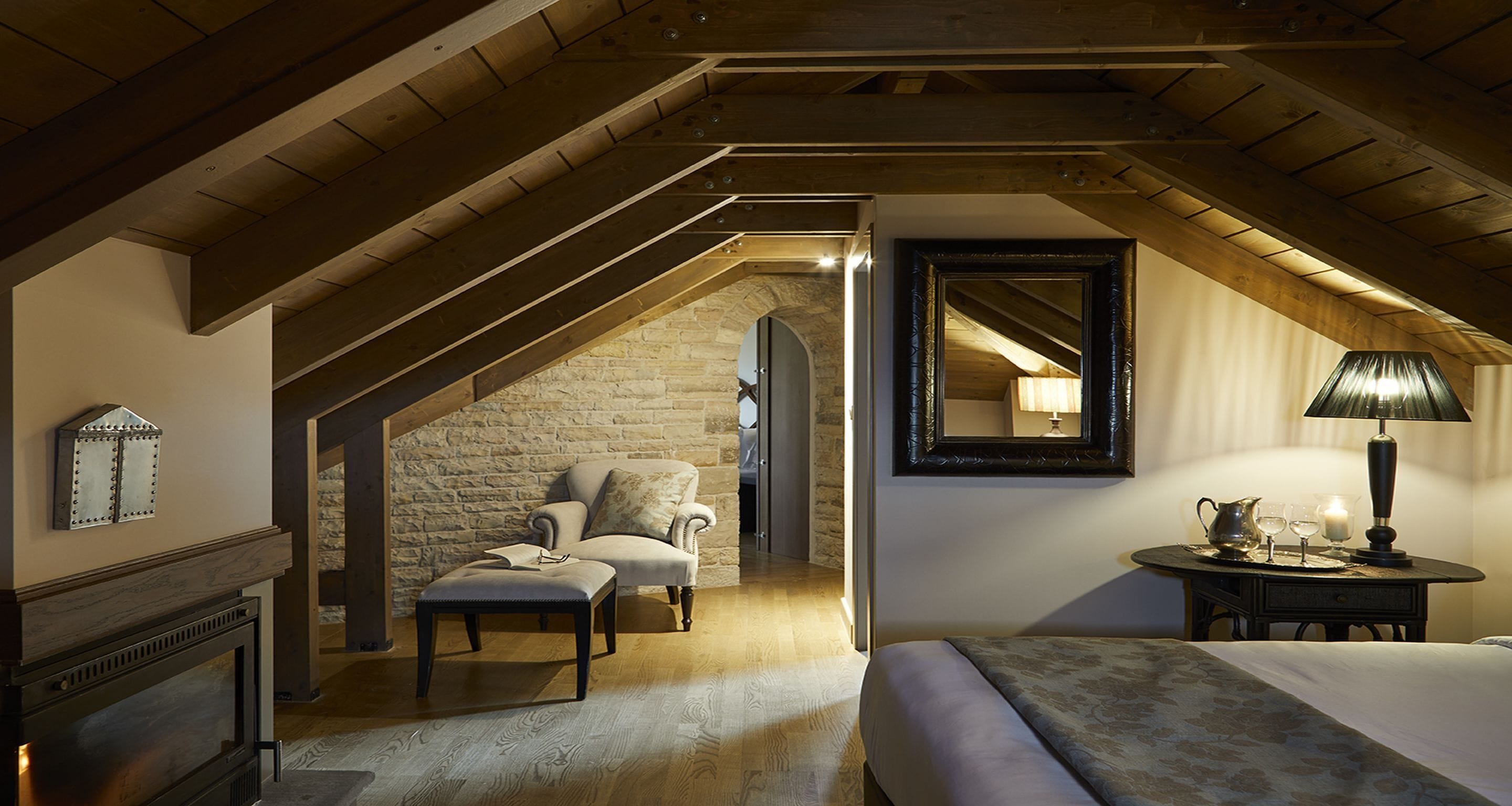 Un dormitorio acogedor en el ático con pared de piedra, chimenea, un sillón cómodo, cama, una mesa con lámpara y espejo.