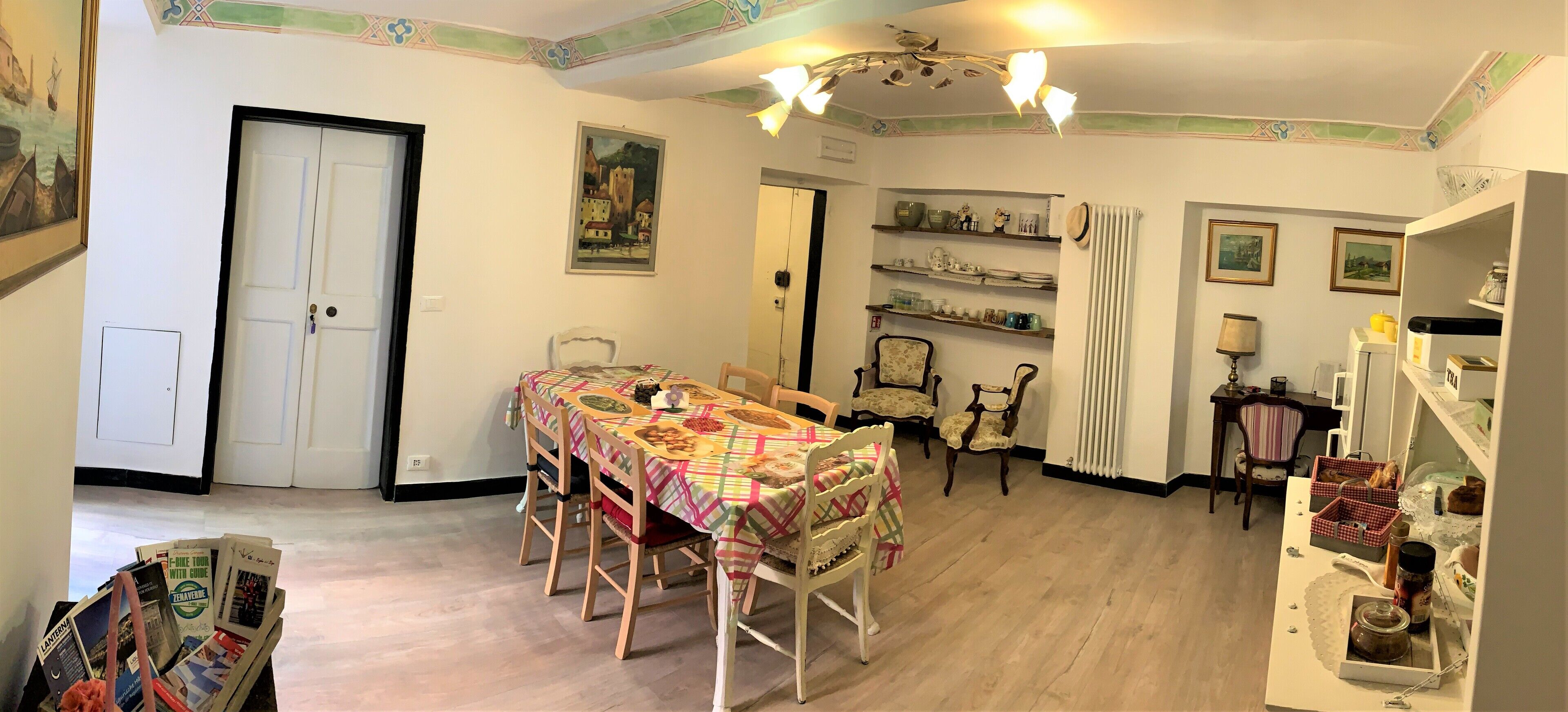 A dining area with a table set for four, surrounded by chairs. There are shelves with dishes and a radiator. A door and a framed picture are visible on the walls.