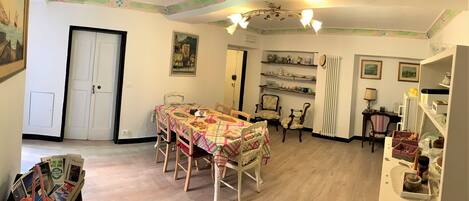 A dining area with a table set for four, surrounded by chairs. There are shelves with dishes and a radiator. A door and a framed picture are visible on the walls.