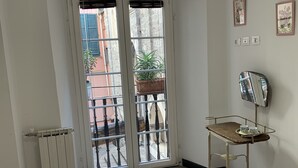 Ein Zimmer mit einem Bett, einem Heizkörper, einer Glastür mit Schiebeöffnung, die auf einen Balkon mit einem Blumentopf führt, einem kleinen Tisch mit Tassen und einem Spiegel.