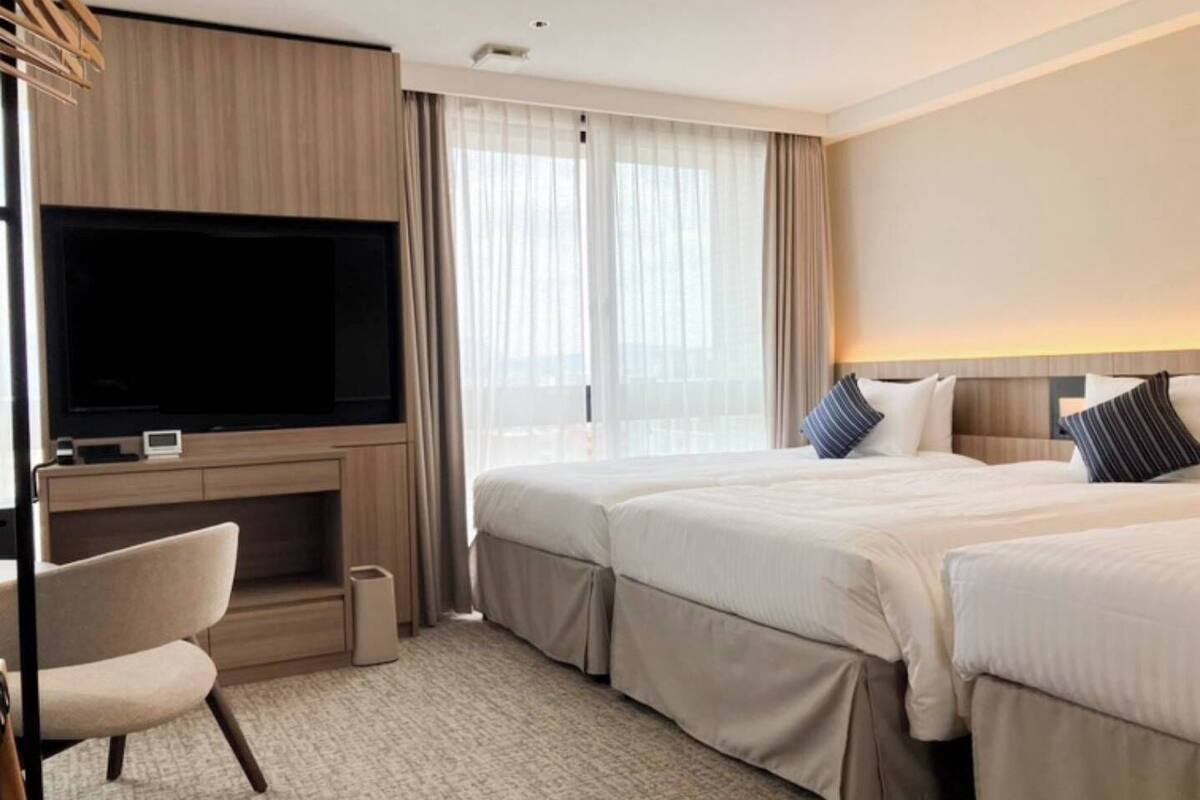 Ein Hotelzimmer mit Fernseher, zwei Betten, einem Schreibtisch und einem Fenster mit Vorhängen.