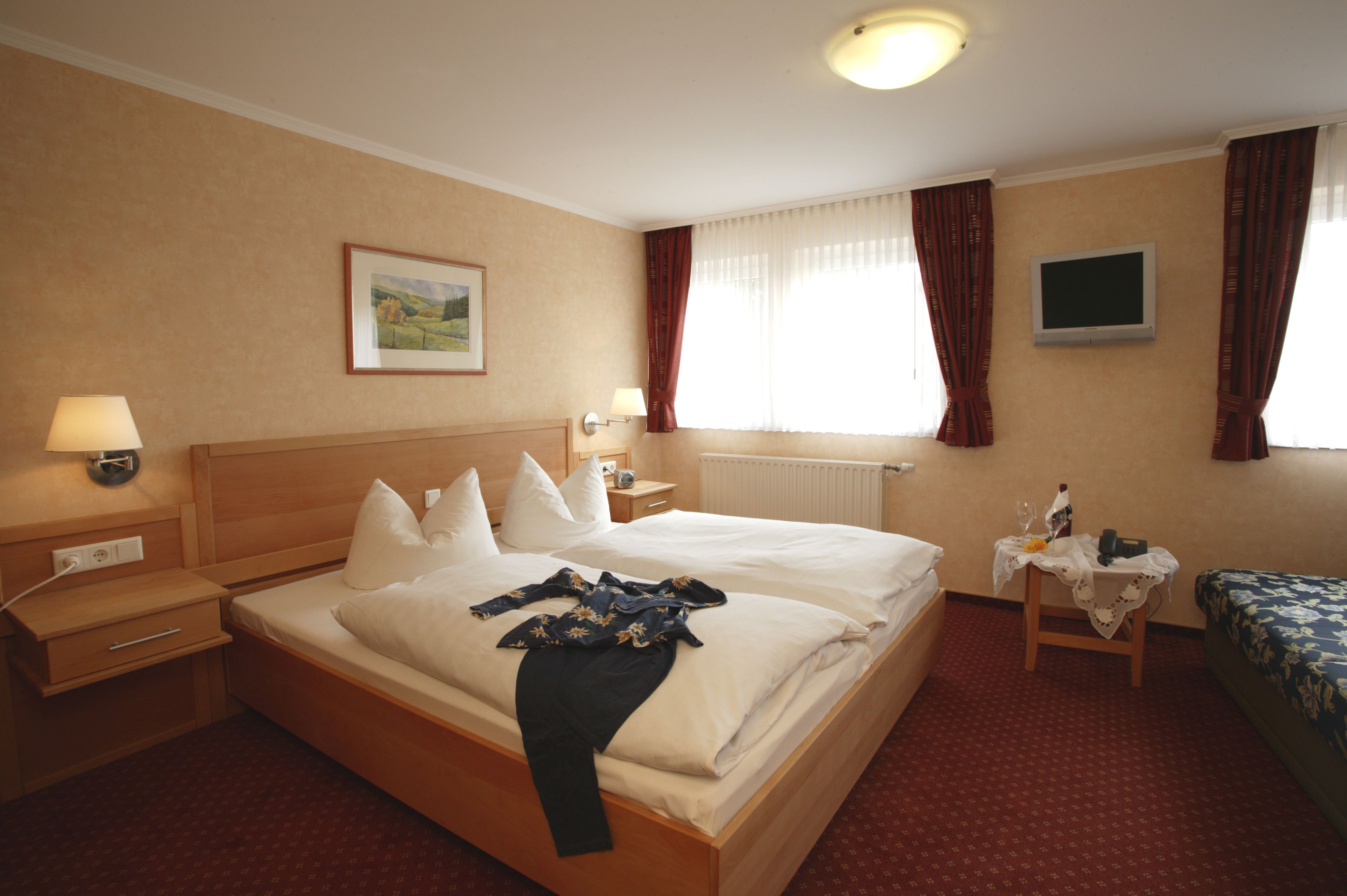 A hotel room with a large bed, a bedside table, a small table with a lamp, a TV, and a window with curtains.