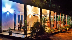 A covered outdoor area with a seating arrangement, potted plants, and a building with illuminated windows.