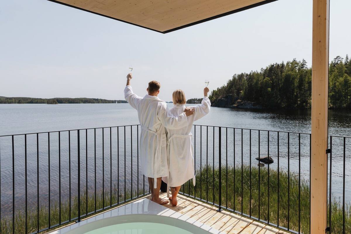 A couple in white robes on a wooden deck overlooking a lake, with a metal railing and a hot tub in the foreground.