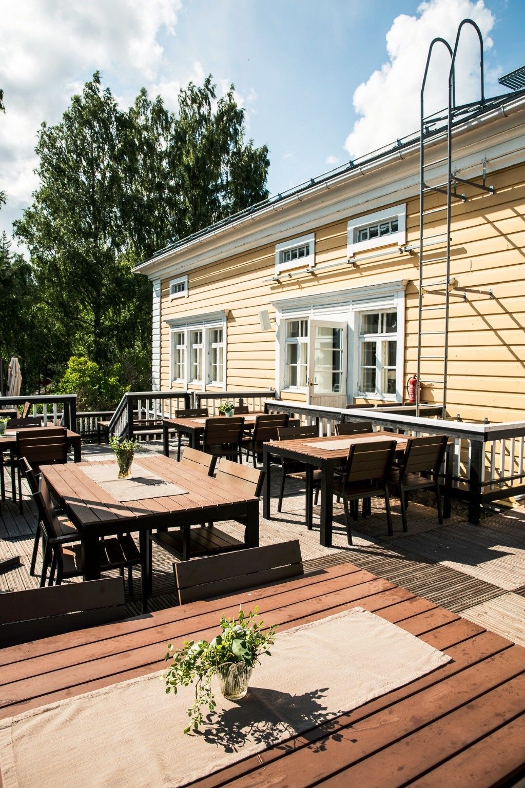 A wooden deck with tables and chairs, a building with windows, and a tree in the background.