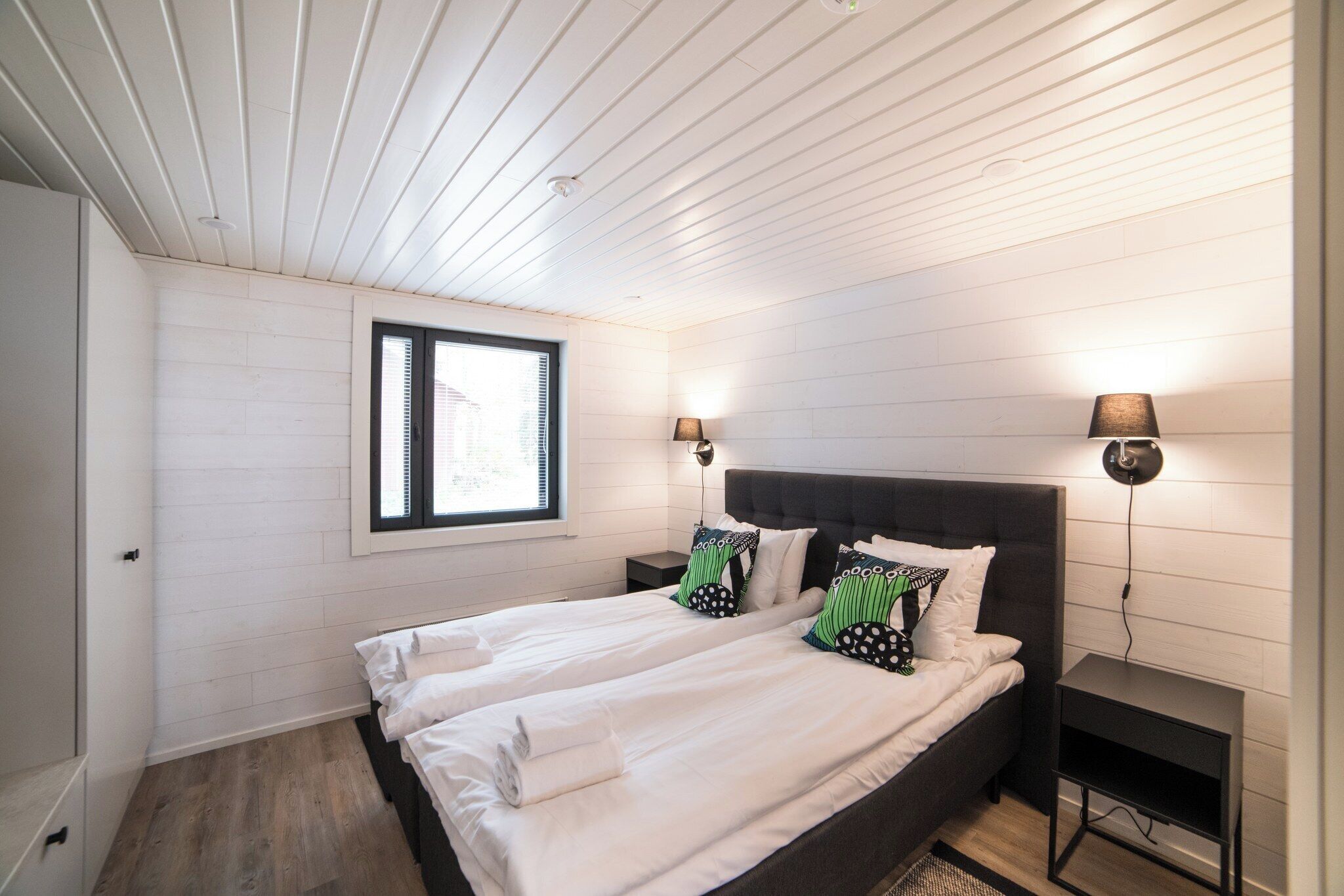 Un dormitorio con dos camas, paredes blancas, techo de madera y una ventana con persianas.
