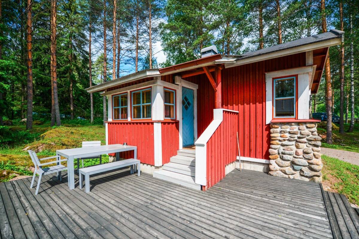 A red wooden cabin with a stone foundation, a deck with a table and benches, and a blue door.
